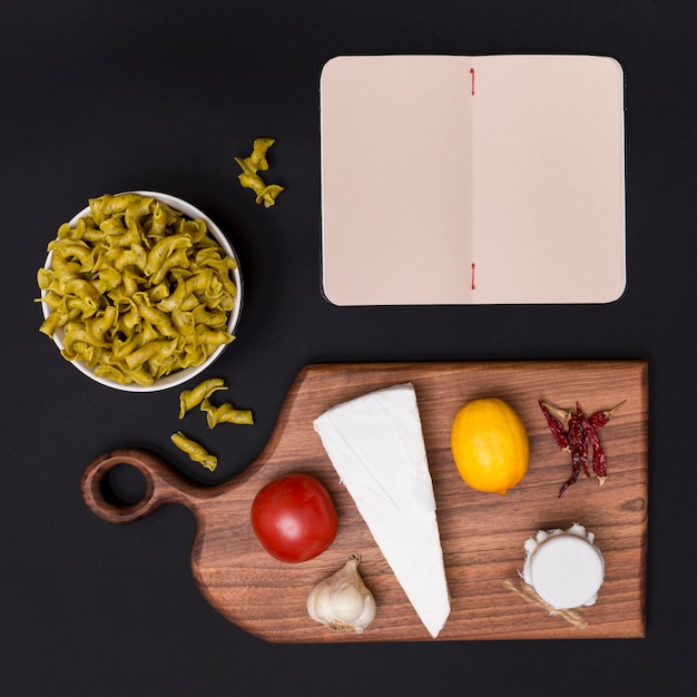 Vista de arriba de las pastas crudas italianas; ingrediente saludable Tabla de cortar y diario en blanco sobre fondo negro