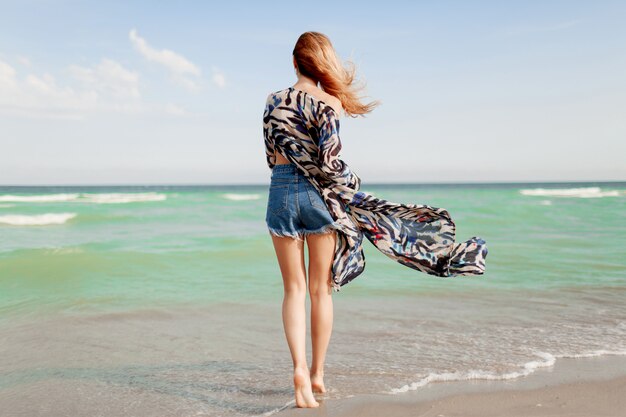 Vista desde atrás de despreocupada mujer elegante con increíbles pelos de jengibre corriendo por la playa