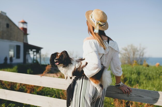 Vista desde atrás en elegante mujer en campo, sosteniendo un perro, feliz estado de ánimo positivo, verano, sombrero de paja, traje de estilo bohemio,