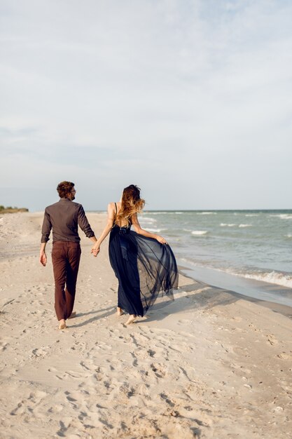 Vista desde atrás. Elegante pareja de enamorados caminando por la playa. Momentos románticos. Olas de arena y mar blancas. Vacaciones tropicales. Altura completa.