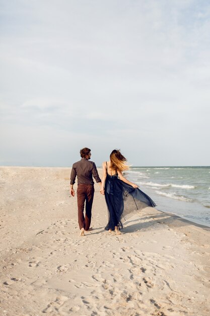 Vista desde atrás. Elegante pareja de enamorados caminando por la playa. Momentos románticos. Olas de arena y mar blancas. Vacaciones tropicales. Altura completa.