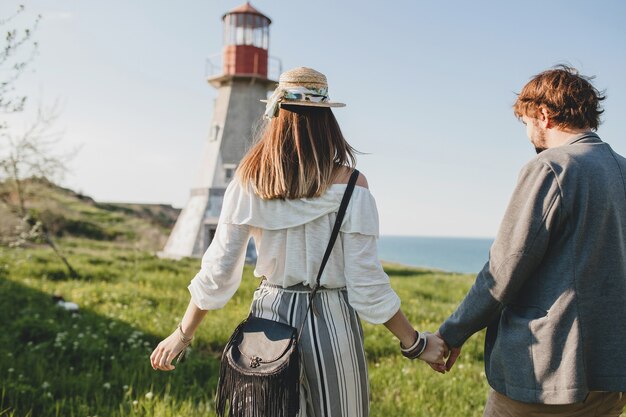 Vista desde atrás en estilo indie hipster pareja joven enamorado caminando en el campo, tomados de la mano, faro en el fondo, día cálido de verano, soleado, traje bohemio, sombrero