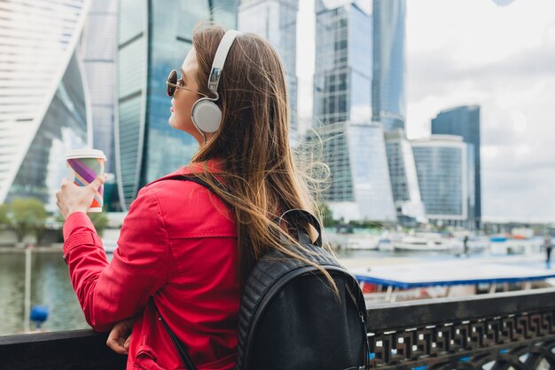 Vista desde atrás de la mujer joven inconformista en abrigo rosa, jeans caminando en la calle con mochila y café escuchando música en auriculares