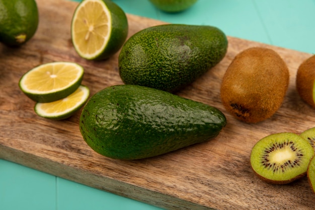 Vista de cerca de aguacates frescos con limas y kiwis aislado en una tabla de cocina de madera sobre un fondo azul.