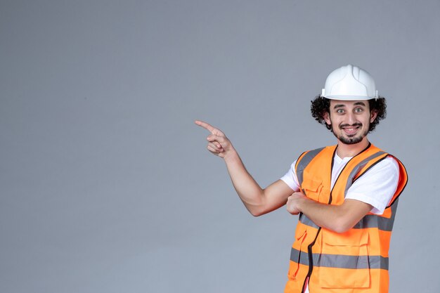 Vista de cerca del arquitecto masculino sonriente en chaleco de advertencia con casco de seguridad sobre pared de onda gris