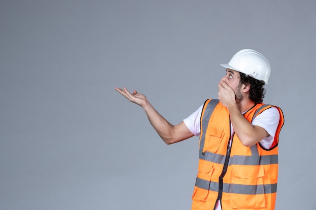 Vista de cerca del arquitecto masculino sorprendido en chaleco de advertencia con casco de seguridad y apuntando algo en el lado derecho en la pared gris de la ola