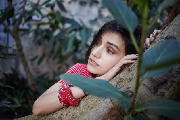 Vista de cerca de la atractiva joven romántica con peinado bob apoyando la cabeza en el tallo del árbol y mirando hacia los lados con expresión facial pensativa de ensueño. Enfoque selectivo en la cara de la niña.