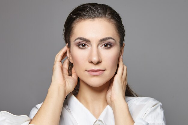 Vista de cerca de la cabeza y los hombros de la elegante y hermosa mujer morena caucásica joven con ojos marrones y maquillaje vistiendo camisa blanca cogidos de la mano en la cara. Moda, glamour y estilo