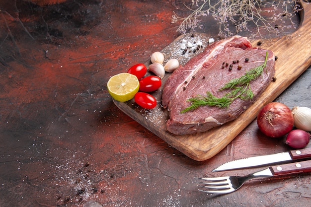 Vista de cerca de la carne roja en la tabla de cortar de madera y ajo, limón, cebolla verde, tenedor y cuchillo sobre fondo oscuro