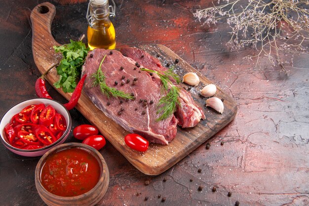 Vista de cerca de la carne roja en la tabla de cortar de madera y ajo verde pimiento picado botella de aceite caído salsa de tomate sobre fondo oscuro