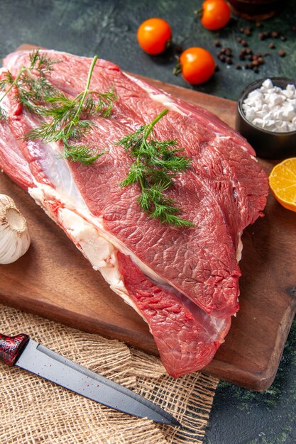 Vista de cerca de carnes rojas crudas frescas ajos verdes sal de limón en un cuchillo de tabla de cortar de madera marrón en una toalla de color nude tomates pimienta sobre fondo de color oscuro