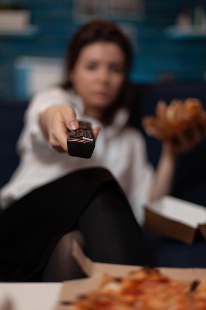 Vista de cerca del control remoto en la mano de la mujer cambiando los canales de televisión mientras cena una hamburguesa de comida rápida para llevar en la sala de estar. Detalle de la persona que navega por las estaciones de televisión sentada en el sofá.