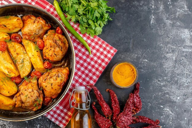 Vista de cerca de una deliciosa cena con pollos, papas, verduras en una cacerola sobre una toalla roja doblada, pimientos secos, botella de aceite caída, paquete verde jengibre amarillo sobre fondo de color oscuro