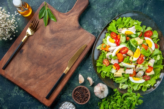 Vista de cerca de una deliciosa ensalada con muchos ingredientes frescos, especias, botella de aceite, ajos, cubiertos, en tabla de cortar de madera