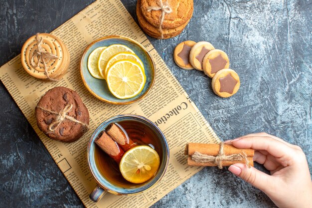 Vista de cerca de deliciosas galletas y mano sosteniendo una taza de té negro con canela en un periódico viejo sobre fondo oscuro