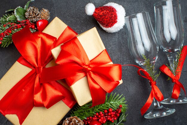 Vista de cerca del estado de ánimo navideño con hermosos regalos con cinta en forma de arco y accesorios de decoración de ramas de abeto sombrero de santa claus copas de cristal conos de coníferas sobre un fondo oscuro