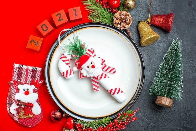 Vista de cerca del fondo de año nuevo con accesorios de decoración de plato de cena ramas de abeto y números calcetín de Navidad en una servilleta roja junto al árbol de Navidad en una mesa negra