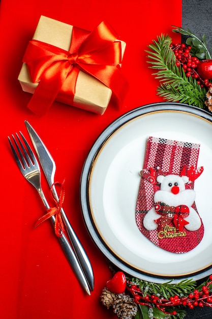 Vista de cerca del fondo de año nuevo con calcetín de Navidad en el plato de cena cubiertos accesorios de decoración ramas de abeto junto a un regalo en una servilleta roja