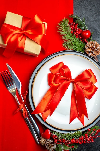 Vista de cerca del fondo de año nuevo con cinta roja en el plato de cena cubiertos accesorios de decoración ramas de abeto junto a un regalo en una servilleta roja