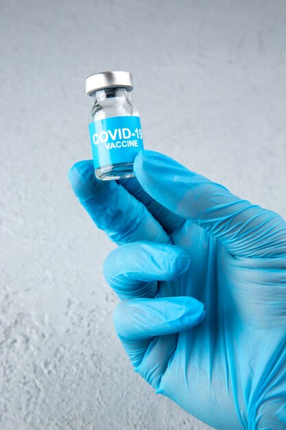 Vista de cerca del guante azul con mano sujetando la ampolla cerrada con vacuna covid- sobre fondo de arena gris