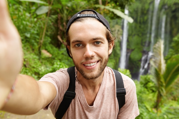 Vista de cerca del guapo excursionista caucásico vistiendo snapback mirando con sonrisa feliz mientras toma selfie con increíble paisaje con cascada