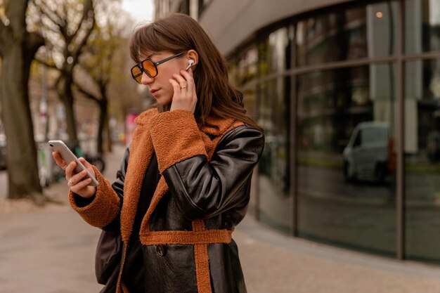 Vista de cerca de una hermosa mujer listando música con gafas de sol en la ciudad mirando el teléfono