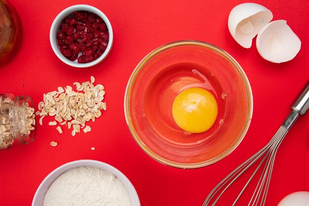 Vista de cerca de los ingredientes como avena de yema de huevo de grosella roja que se derrama de mermelada de harina de tarro para preparar el desayuno con batidor y cáscara de huevo sobre fondo rojo