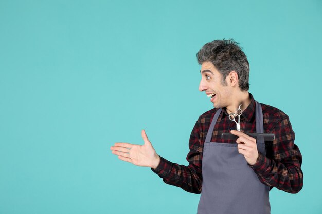 Vista de cerca del joven barbero sorprendido con delantal gris y sosteniendo el peine y la tijera apuntando algo en el lado derecho sobre fondo de color azul