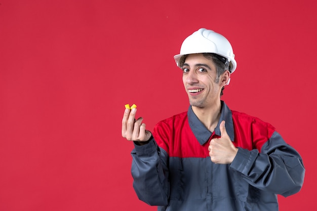 Foto gratuita vista de cerca del joven constructor confiado en uniforme con casco y sosteniendo tapones para los oídos haciendo un gesto aceptable en la pared roja aislada