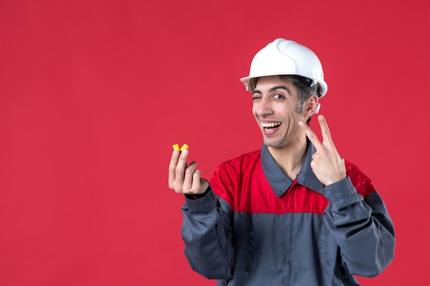 Foto gratuita vista de cerca del joven constructor confiado en uniforme con casco y sosteniendo tapones para los oídos haciendo gesto de victoria en la pared roja aislada