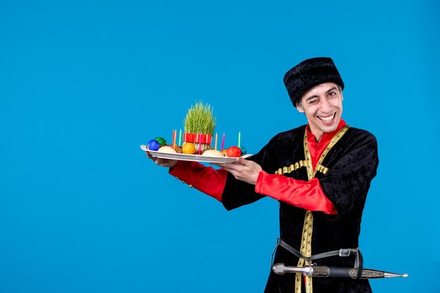 Vista de cerca de un joven emotivo y divertido vestido con un traje tradicional que da una bandeja llena de dulces nacionales de fondo azul