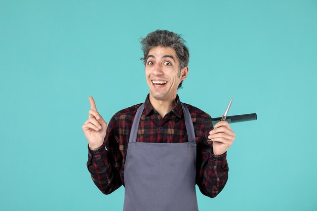 Vista de cerca del joven peluquero masculino sonriente vistiendo delantal gris y sosteniendo el peine de tijera apuntando hacia arriba sobre fondo de color azul pastel