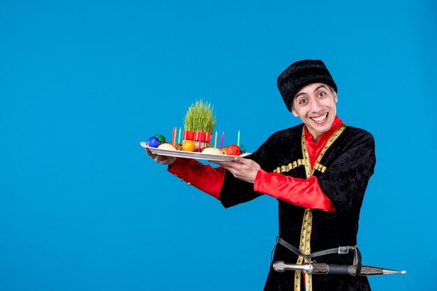 Vista de cerca de un joven sonriente y confiado vestido con un traje tradicional que da una bandeja llena de confitería nacional con fondo azul