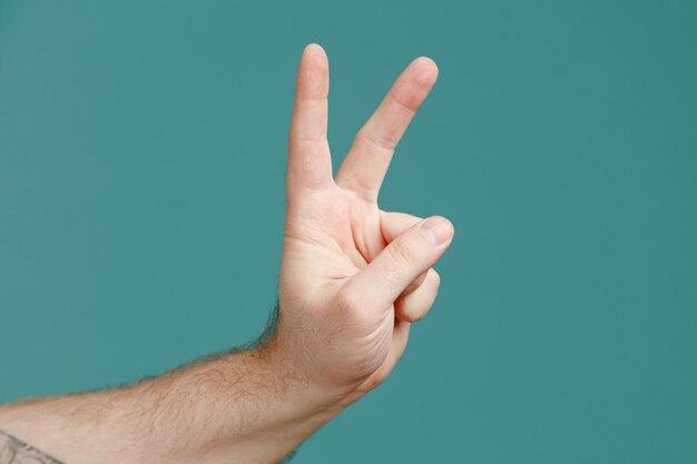 Vista de cerca de la mano masculina joven que muestra el signo de la paz aislado sobre fondo azul