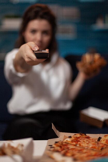 Vista de cerca de la mano de la mujer sosteniendo el control remoto de la televisión para cambiar el volumen mientras cena pizza de entrega en la sala de estar. Detalle de la persona que navega por los canales de televisión mientras come una hamburguesa.