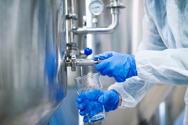 Foto gratuita vista de cerca de las manos en guantes protectores abriendo la válvula del grifo y llenando el vaso con muestras del depósito de la máquina industrial