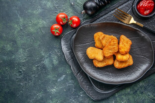 Vista de cerca de nuggets de pollo en un plato negro y salsa de tomate de tenedor elegante en la bandeja de color oscuro tomates de flores blancas en el lado izquierdo