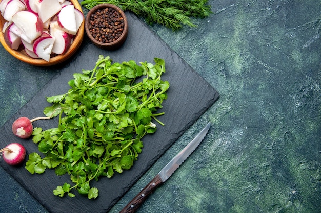 Vista de cerca del paquete de cilantro pimiento de rábanos picados enteros frescos en una tabla de cortar de madera y un cuchillo en el lado derecho sobre fondo verde negro de colores mezclados con espacio libre