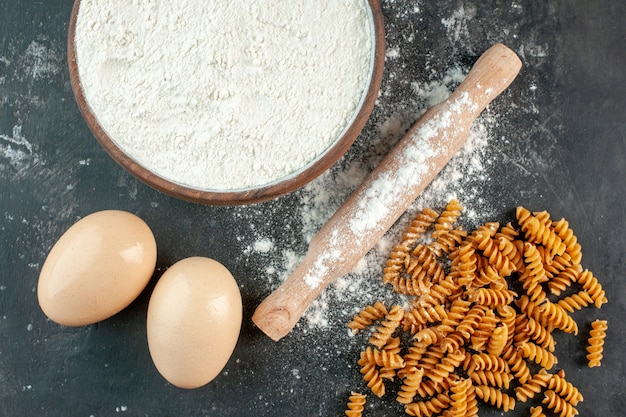 Vista de cerca de pastas italianas crudas dos huevos y harina en un cuenco marrón con rodillo sobre fondo gris