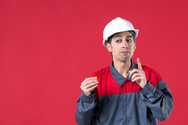 Vista de cerca del pensamiento joven trabajador en uniforme con casco y sosteniendo tapones para los oídos en la pared roja aislada