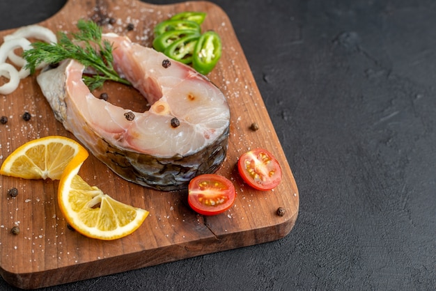 Vista de cerca de pescado crudo y verduras frescas picadas rodajas de limón especias sobre una tabla de madera en el lado derecho sobre la superficie angustiada negra