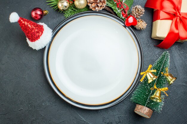 Vista de cerca del plato de cena árbol de navidad ramas de abeto cono de coníferas caja de regalo sombrero de santa claus sobre fondo negro