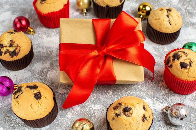 Vista de cerca del regalo con cinta roja entre deliciosos cupcakes pequeños recién horneados y accesorios de decoración en la mesa de hielo