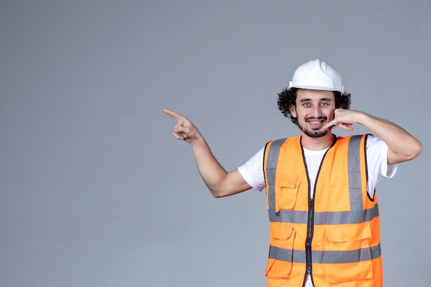 Vista de cerca del sonriente arquitecto masculino en chaleco de advertencia con casco de seguridad y apuntando algo en el lado derecho haciendo un gesto de llamarme en la pared de onda gris