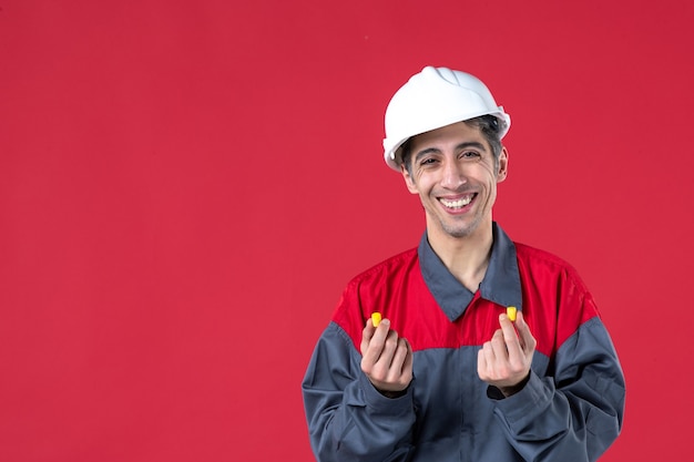 Vista de cerca del sonriente joven constructor en uniforme con casco y sosteniendo tapones para los oídos en la pared roja aislada