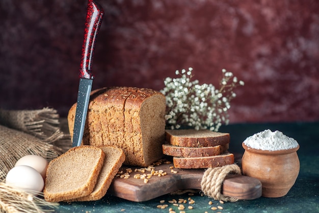 Vista de cerca de los trigos de pan negro dietéticos en la tabla de cortar de madera cuchillo flor huevos harina en un tazón toalla marrón sobre fondo de colores mezclados