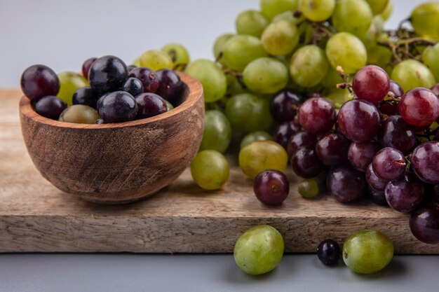 Vista cercana de bayas de uva en un tazón y uvas en la tabla de cortar sobre fondo gris