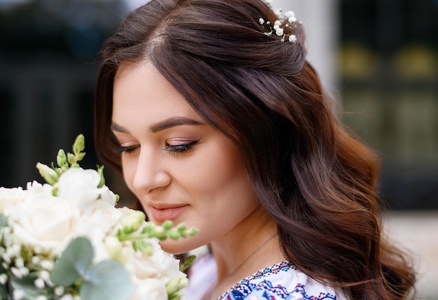 Vista cercana de la cara encantadora de una joven que tiene el pelo rizado moreno y un lindo maquillaje sosteniendo flores de novia y mirando hacia abajo mientras lo huele