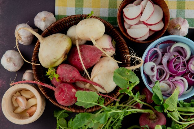 Vista cercana de la cesta y cuencos llenos de verduras como cebolla de rábano y ajo sobre fondo marrón