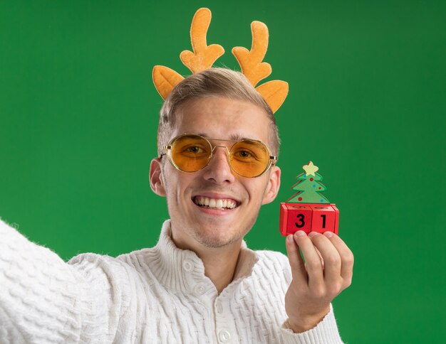 Foto gratuita vista cercana del chico guapo joven sonriente con diadema de cuernos de reno con gafas sosteniendo un juguete de árbol de navidad con fecha extendiendo la mano hacia la cámara mirando aislado en la pared verde